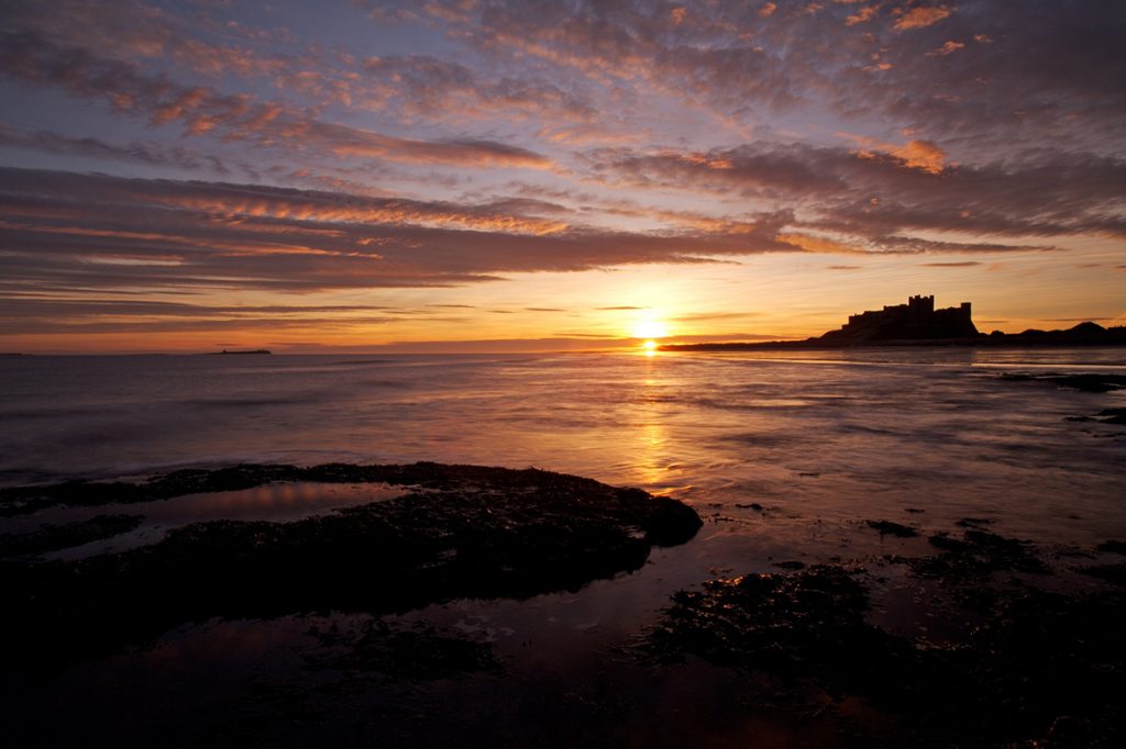 Mike Caine - Stocklandscapes - Stock Photography Bamburgh Castle, Northumberland; sunrise. Wide across sea and rocks